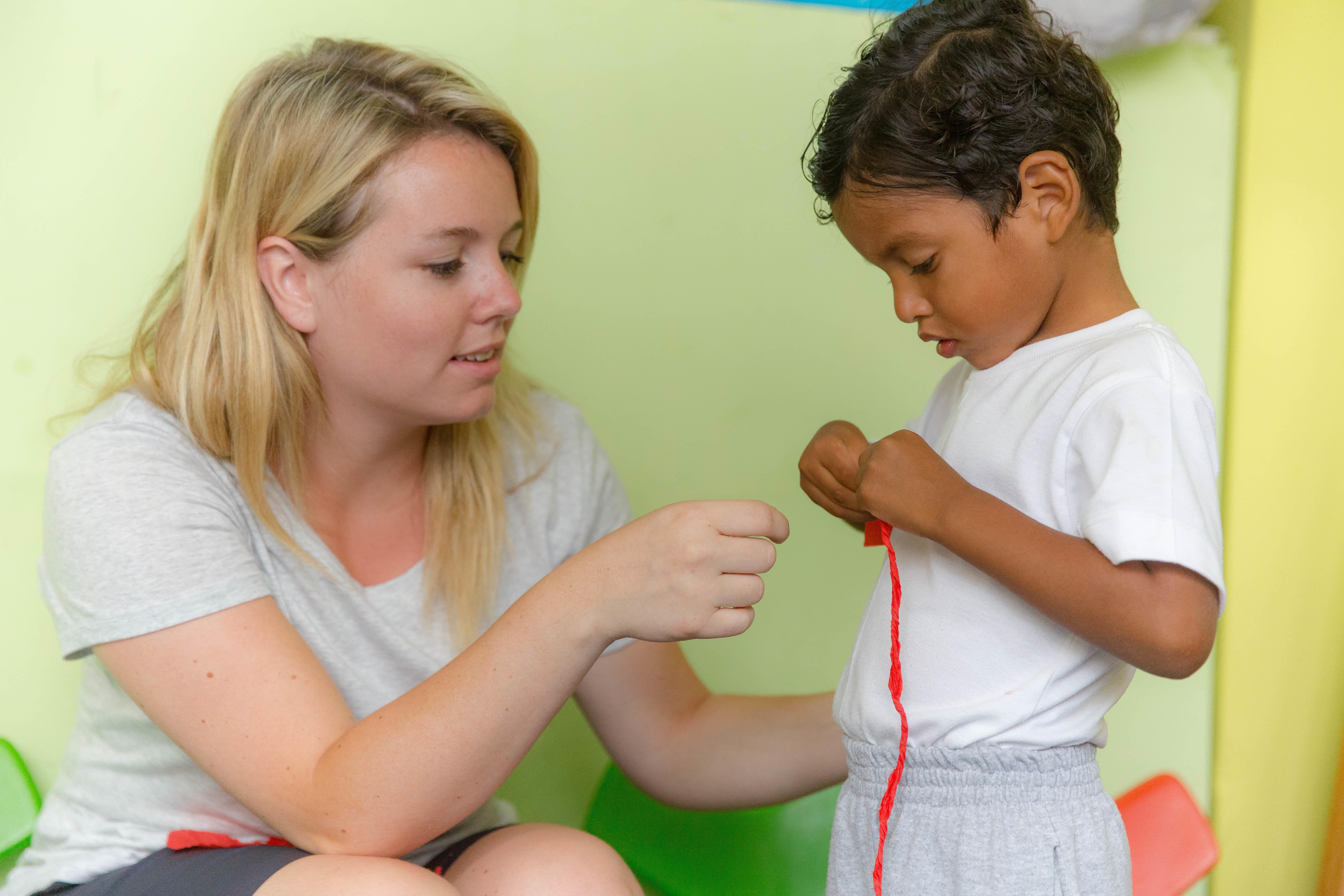 Female Childcare volunteer helps a young child with a ribbon during an arts and craft activity at a kindergarten in Ecuador. Projects Abroad volunteer working with children in Ecuador helps child with an activity in a day care centre.